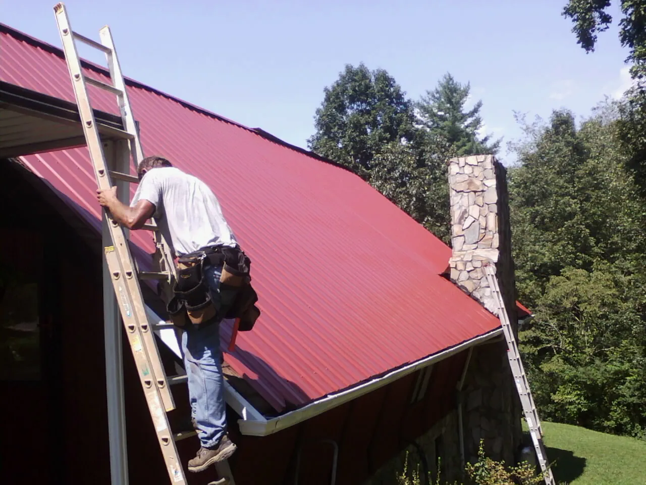 Roofer on a ladder beside a red metal roof and stone chimney — Soffit & Fascia Repair by Roofing Brothers in Summerton